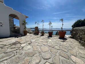 A stone-paved terrace with potted plants, a white building with an archway, and a view of the sea.