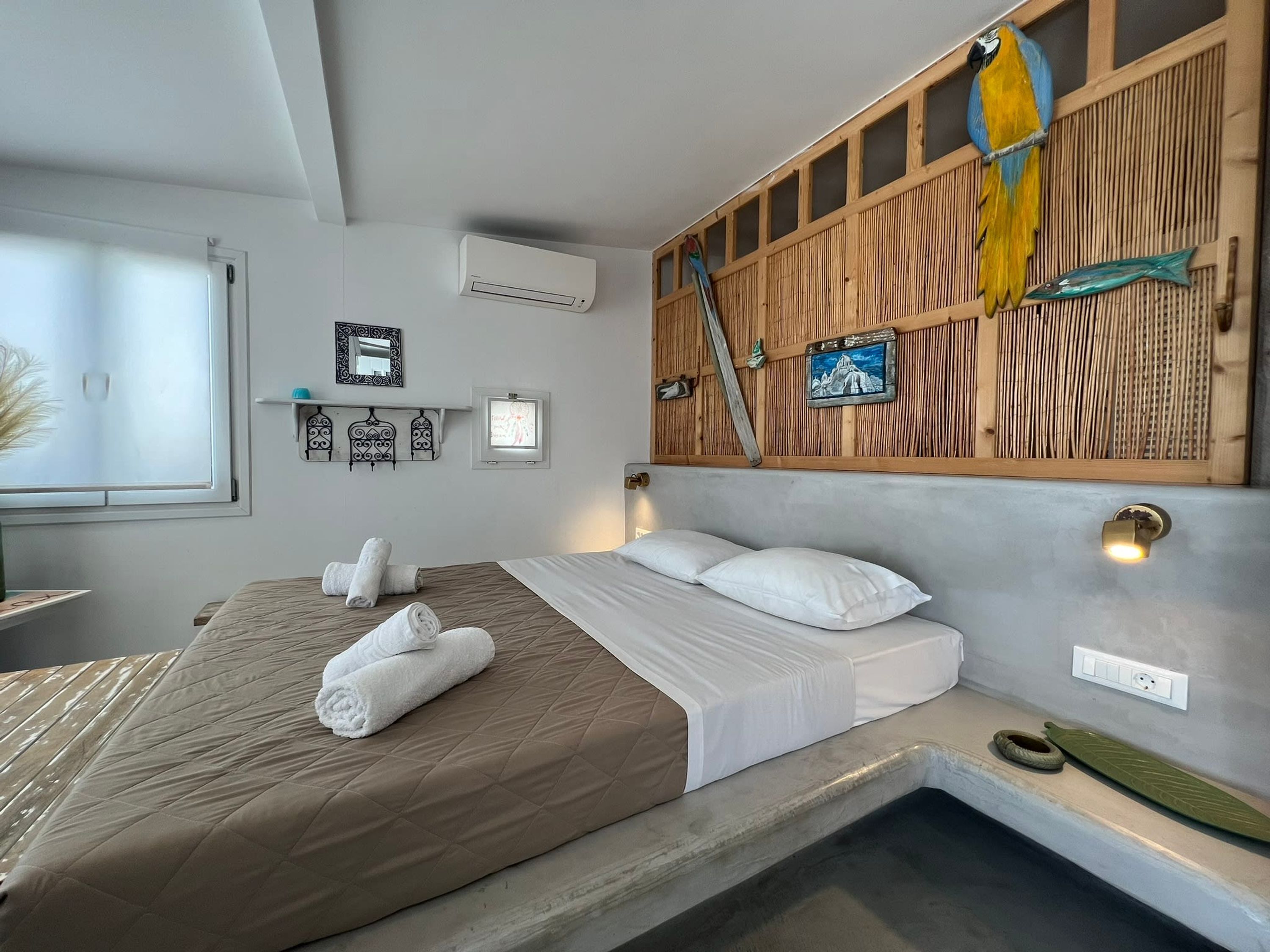 Une chambre d’hôtel comprenant un lit, une petite table de chevet, un climatiseur fixé au mur, un panneau en bois orné d’un motif d’oiseau et un tableau encadré.