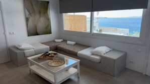 A modern, minimalist living room with a large window offering a view of the sea. There is a glass coffee table with a tray of pastries and a cup of coffee. - Mykonos Noir Nest - Sea View Escape (Mykonos)