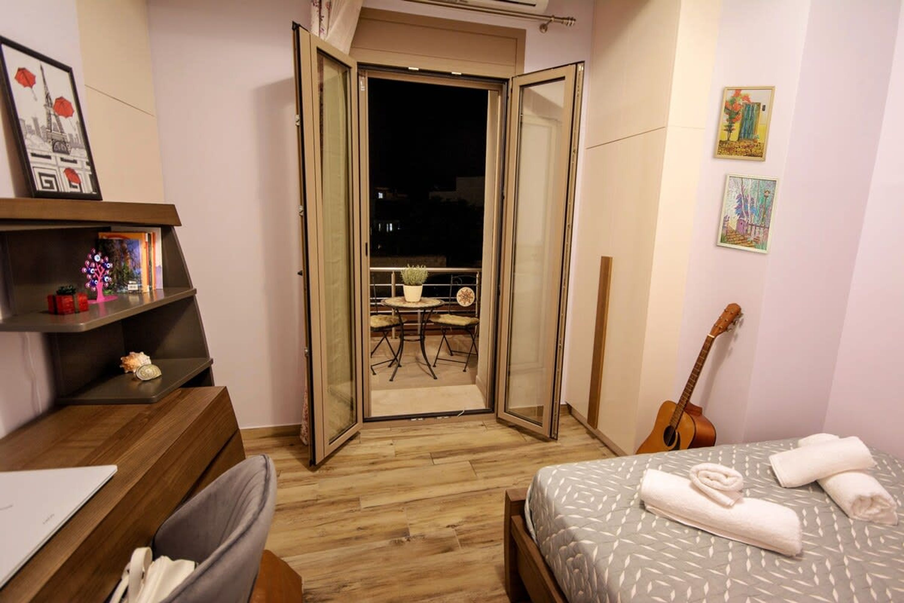 Et soveværelse med seng, skrivebord, en guitar og en balkon med bord og stole.
