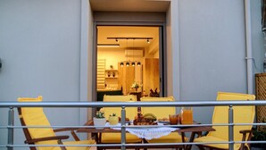 Ένας εξωτερικός χώρος για φαγητό με κίτρινες καρέκλες και ένα τραπέζι, ένα προστατευτικό κιγκλίδωμα και θέα σε έναν εσωτερικό χώρο μ ε ζεστό φωτισμό και έπιπλα από ξύλο.