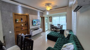 A modern living room with a TV, green sofa, wooden shelves, and a dining area with chairs.
