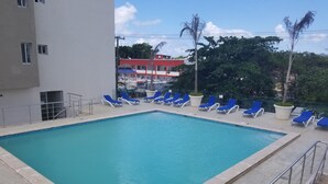 A rectangular swimming pool with blue water, surrounded by a tiled deck. There are blue lounge chairs along the pool's edge, and a metal railing separates the pool from the deck. In the background, there are buildings with red and white facades, and a tree with a nest.