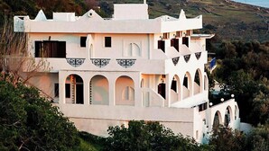 Un edificio blanco con múltiples arcos y balcones, rodeado de vegetación y colinas.
