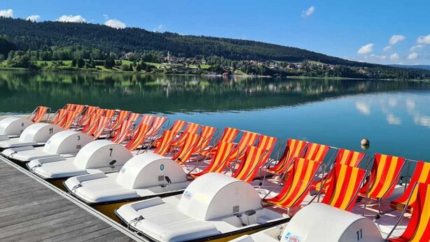 A row of sun loungers with white canopies and red and yellow striped towels, facing a calm lake with a forested hill in the background. - Escape to a Charming Villa in Haut-Doubs. (Bians-les-Usiers)