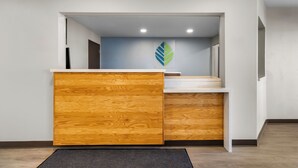 A reception area with a wooden counter, a blue wall with a leaf logo, and a staircase leading to an upper floor. - Woodspring Suites Palmdale - Lancaster (Palmdale)