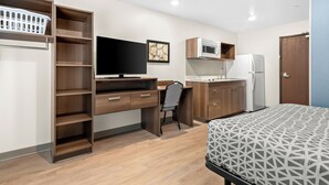 A compact room with a bed, a desk with a chair, a television, a microwave, a refrigerator, and a laundry basket. - Woodspring Suites Palmdale - Lancaster (Palmdale)