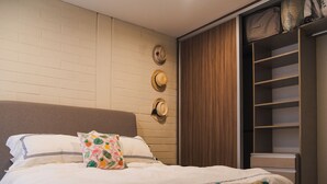 Una cama bien tendida con ropa de cama blanca y un cojín floral, un cabecero de madera, y un armario con espejo y repisas.