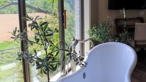 Ein Badezimmer mit Badewanne, einem Fenster mit Blick auf Bäume und einer Topfpflanze auf einem Holzschemel.