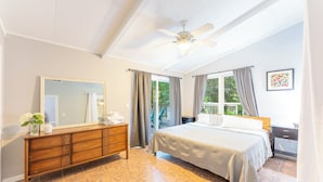 Ein Schlafzimmer mit Bett, Kommode, Deckenventilator und großem Fenster mit Vorhängen.