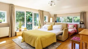 Un dormitorio amplio con una cama grande, un sofá y un escritorio. Varias ventanas ofrecen luz natural y vistas al verdor exterior.