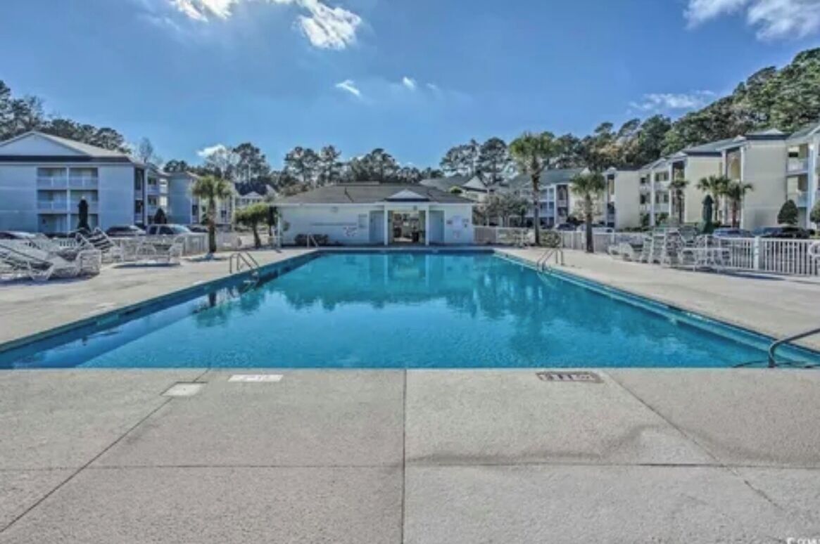 Una piscina rectangular con solárium alicatado, rodeada por una valla, y adyacente a un edificio de techo plano y múltiples ventanas.
