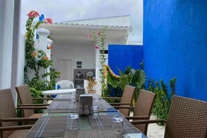An outdoor dining area with wicker chairs and a glass table, surrounded by plants and a blue wall. - Coral Castle Guest House (Goidhoo)