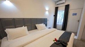 A hotel room with a large bed, a bedside table, a nightstand, a window with curtains, and an air conditioning unit.