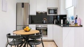 Una cocina moderna con armarios blancos, electrodomésticos de acero inoxidable, una mesa redonda de madera con sillas y una ventana que proporciona luz natural.