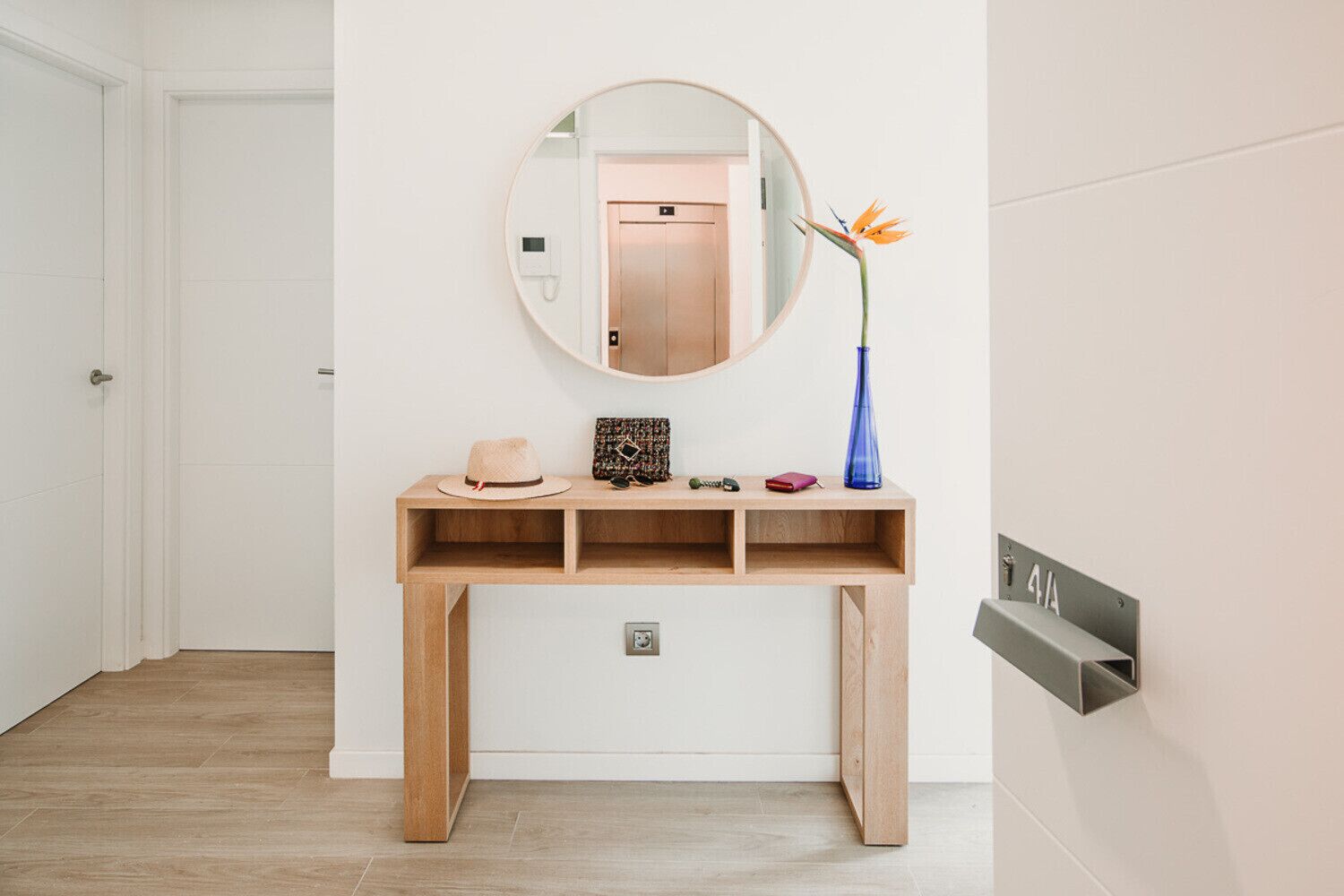 Un baño minimalista con tocador de madera, un espejo redondo y un florero con una sola flor.