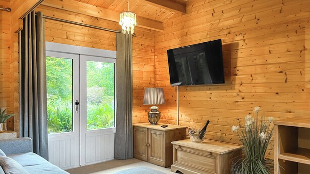 A cozy log cabin room with a sofa, a TV mounted on the wall, a lamp, and a window with curtains. - 1 bedroom accommodation in Rudyard, near Leek (Rudyard, near Leek)