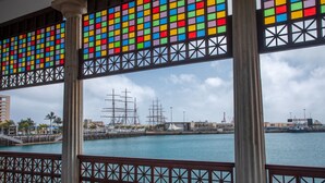 Blick durch ein buntes Fenster mit Geländer auf einen Hafen mit Schiffen und eine Stadtansicht.