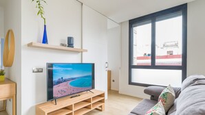 A modern living room with a flat-screen TV on a wooden stand, a grey sofa, a large window with a view of buildings, and a small wooden table with a plant. - VIVENTE 3A BY CANARIAS GETAWAY (Las Palmas de Gran Canaria)
