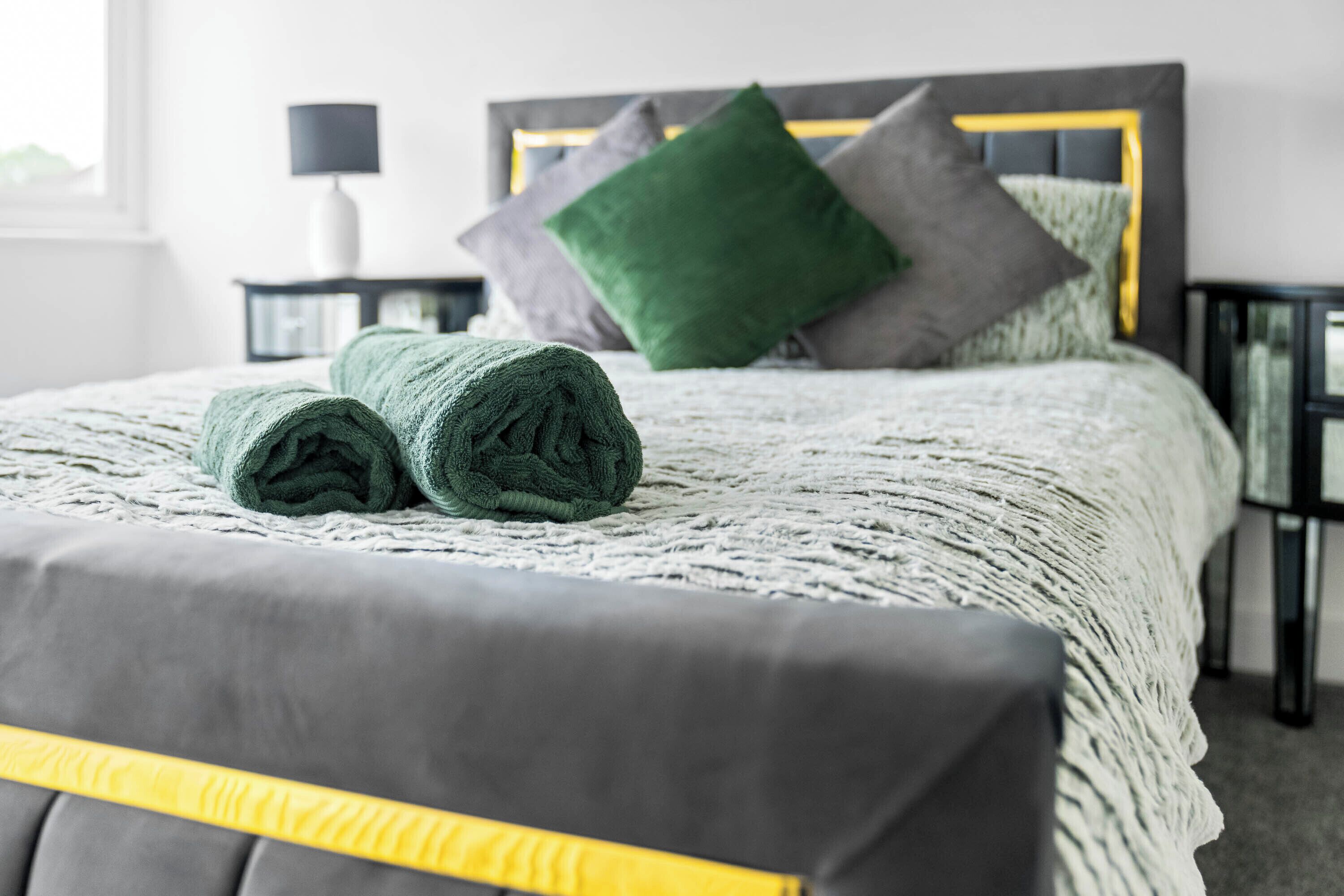 A neatly made bed with a grey and yellow headboard, green and grey pillows, and a white bedside table with a lamp.