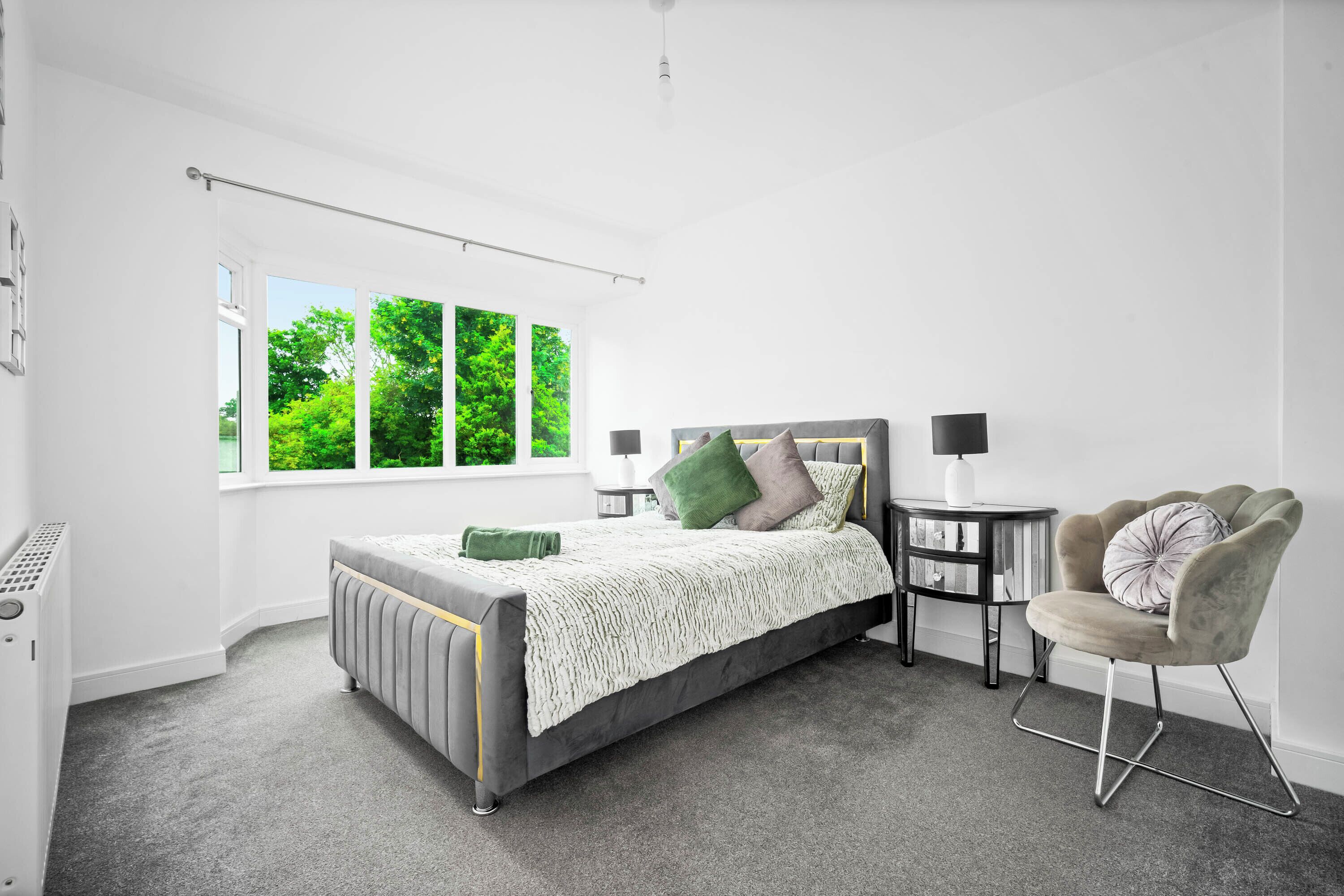 Un dormitorio moderno con un ventanal grande, una cama con cabecero gris, una mesita de noche y una silla.