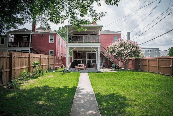 A two-story house with a front porch, a wooden fence, and a small garden with a tree and a hammock.