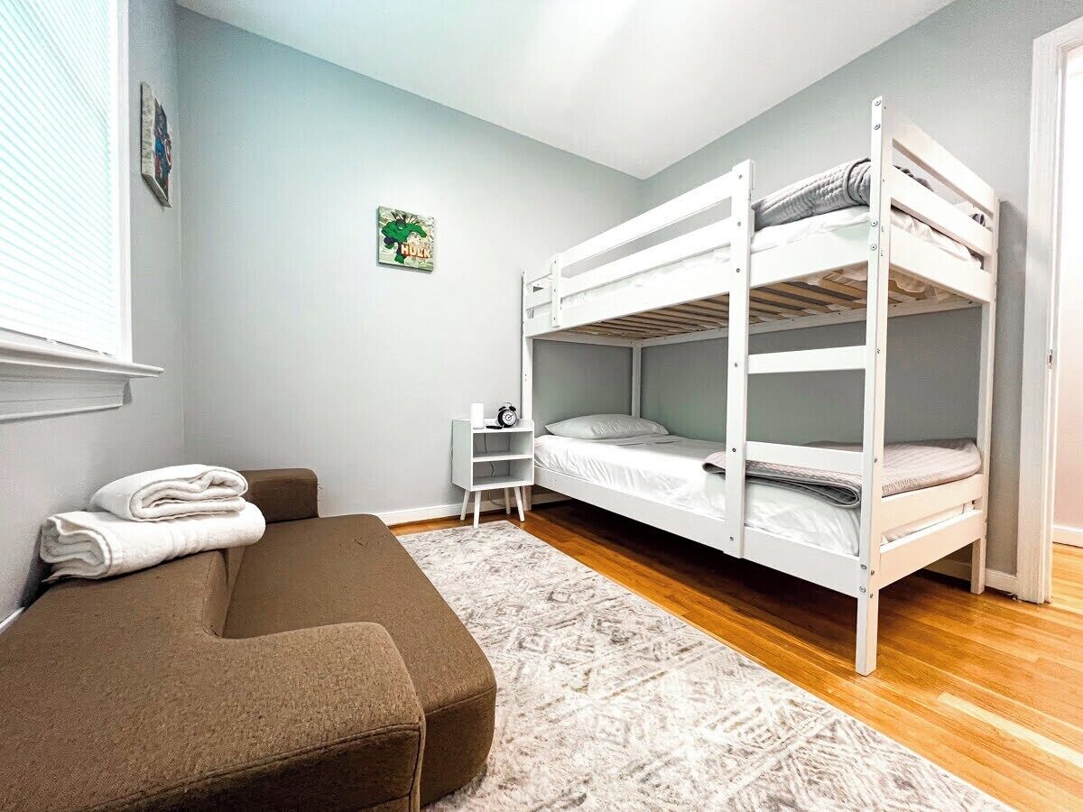 Een kamer met een stapelbed, een bruine bank, een klein wit nachtkastje en een kamerkleed met dessin.