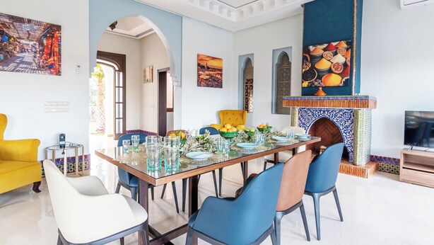 A dining area with a long table set for six, surrounded by blue and white chairs. There is a yellow armchair and a white armchair. A fireplace with a blue and white tiled mantle is visible. - Domaine Mandar Atlas (Marrakech)