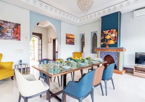 A dining area with a long table set for six, surrounded by blue and white chairs. There is a yellow armchair and a white armchair. A fireplace with a blue and white tiled mantle is visible. - Domaine Mandar Atlas (Marrakech)