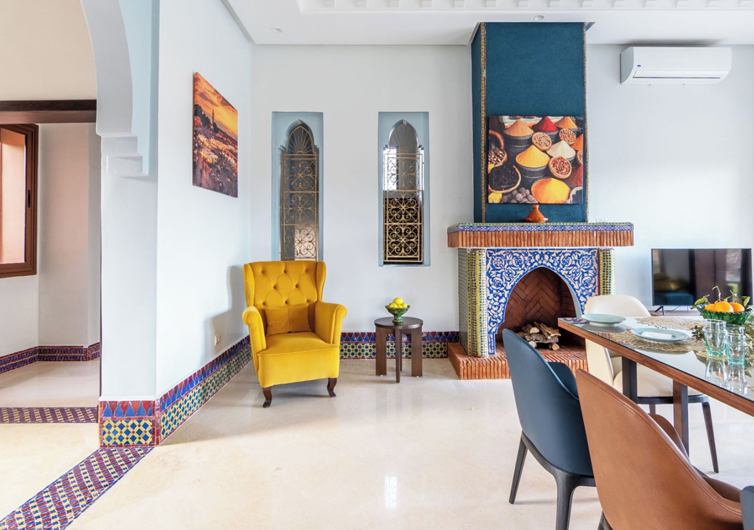 Un salon moderne avec un fauteuil jaune, une table à manger dressée avec des assiettes et des verres, une cheminée bleue à motifs et des carreaux décoratifs le long du sol.