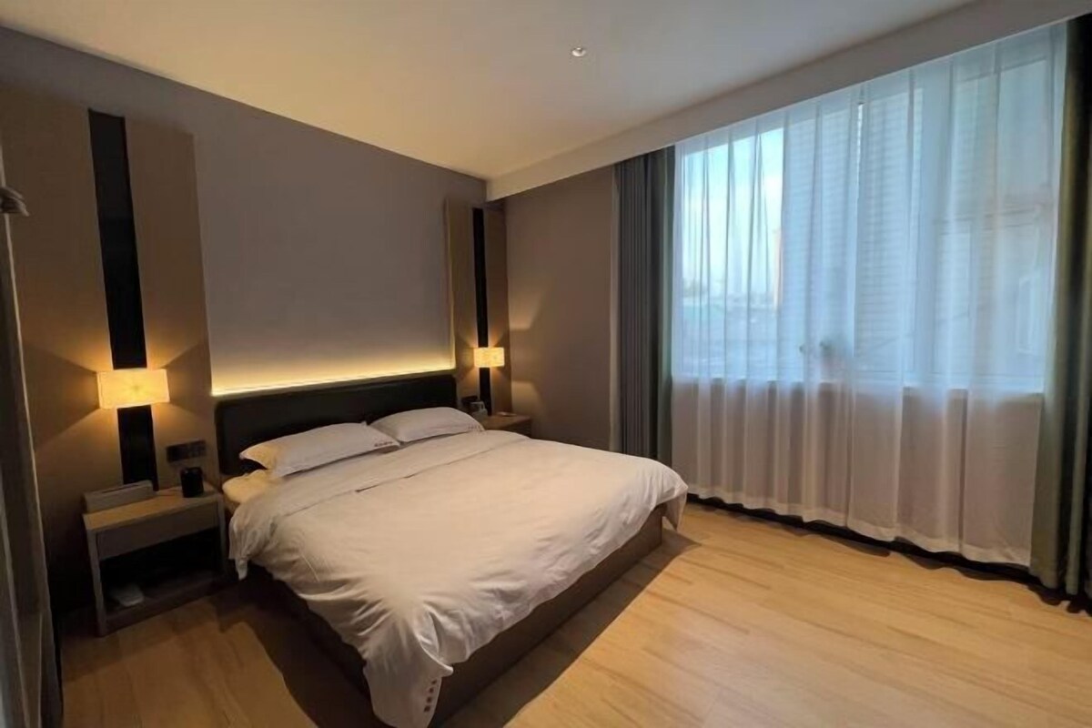 A modern hotel room with a large bed, bedside lamps, a nightstand, and a view of the outdoors through sheer curtains.