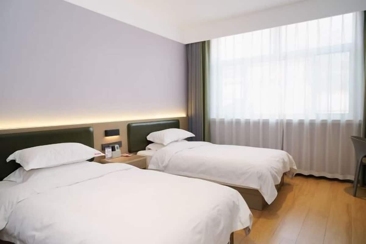 A hotel room with two beds, a nightstand with a lamp, a window with curtains, and a wooden floor.