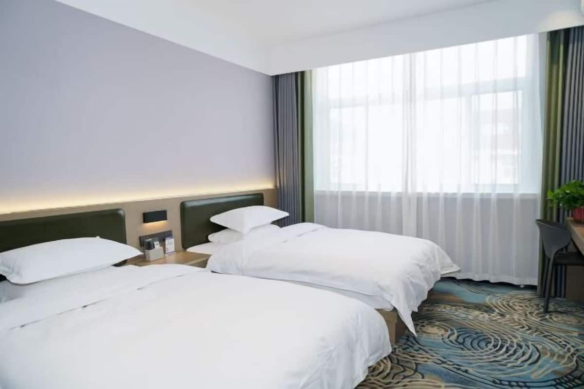A hotel room with two beds, a desk, a chair, a potted plant, and a large window with curtains.
