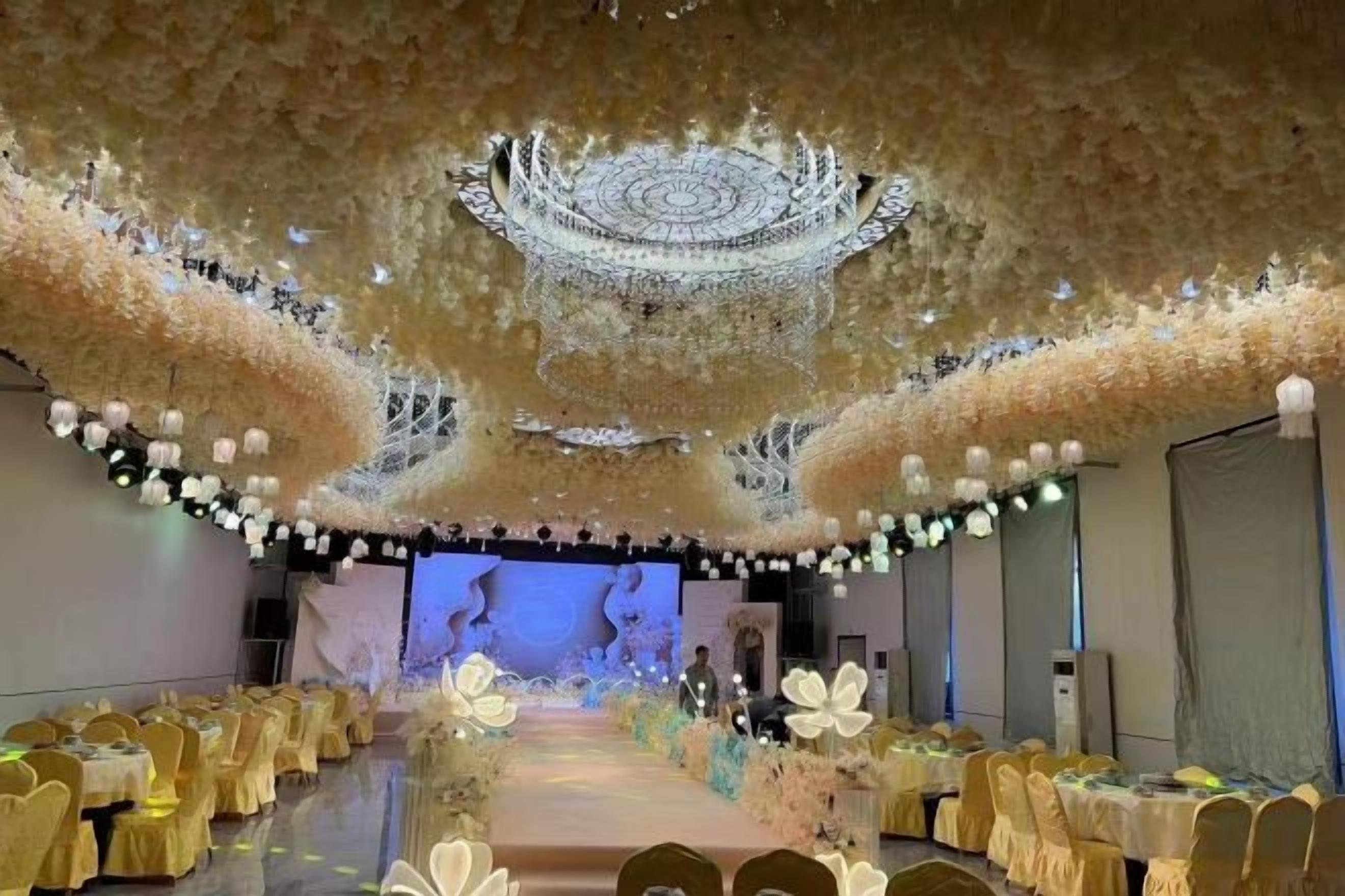 A banquet hall with a stage, rows of tables with yellow tablecloths, and a floral ceiling design.