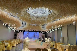 A banquet hall with a stage, rows of tables with yellow tablecloths, and a floral ceiling design.