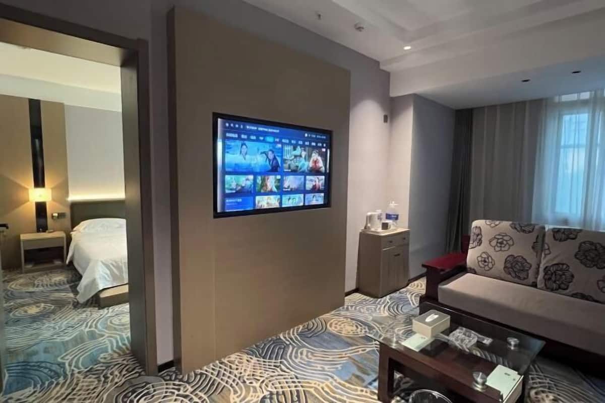 A hotel room with a large TV mounted on the wall displaying a video, a sofa, a coffee table, a bedside table, and a patterned carpet.