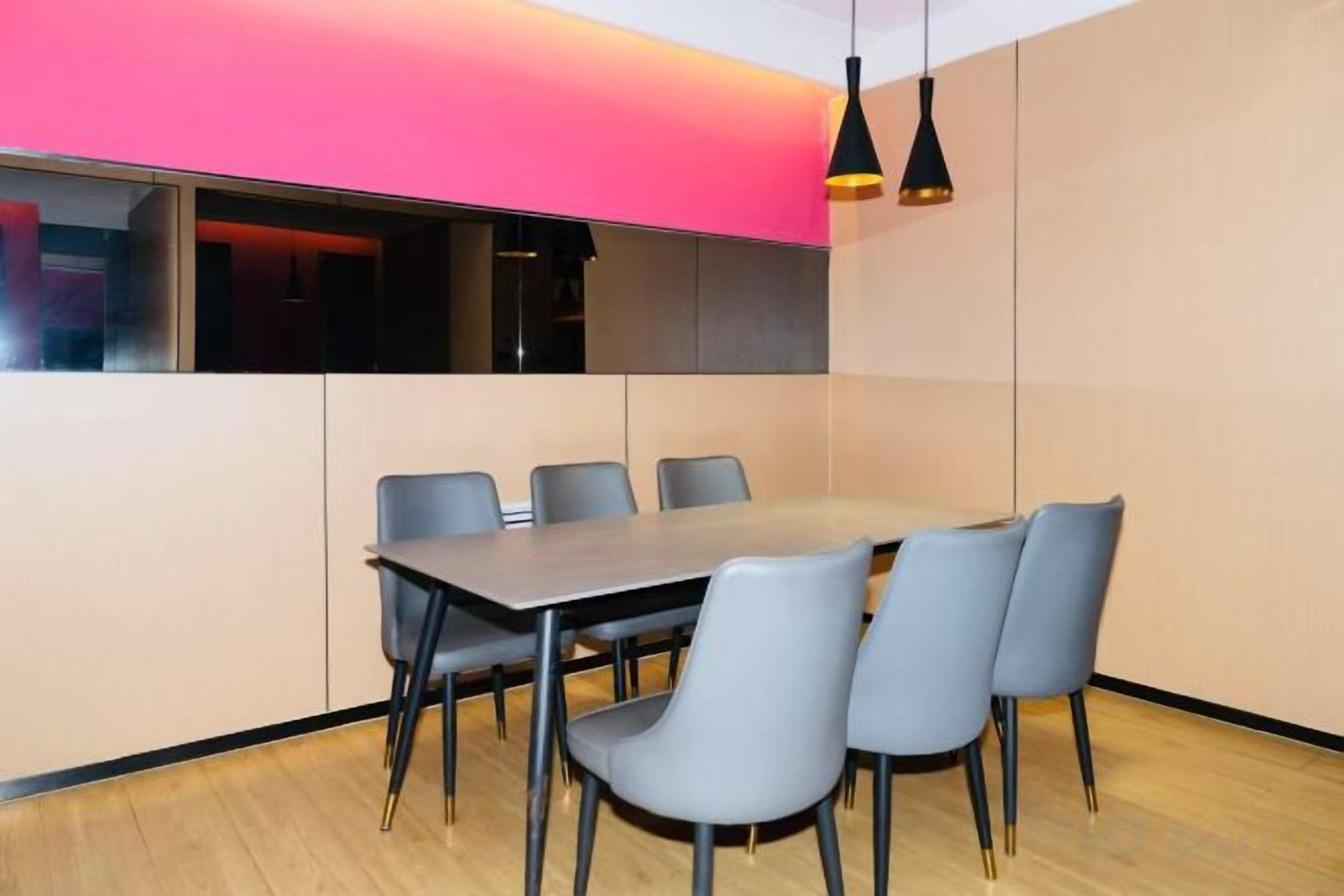 A conference room with a long table and six chairs, a pink accent wall, and a black pendant light.