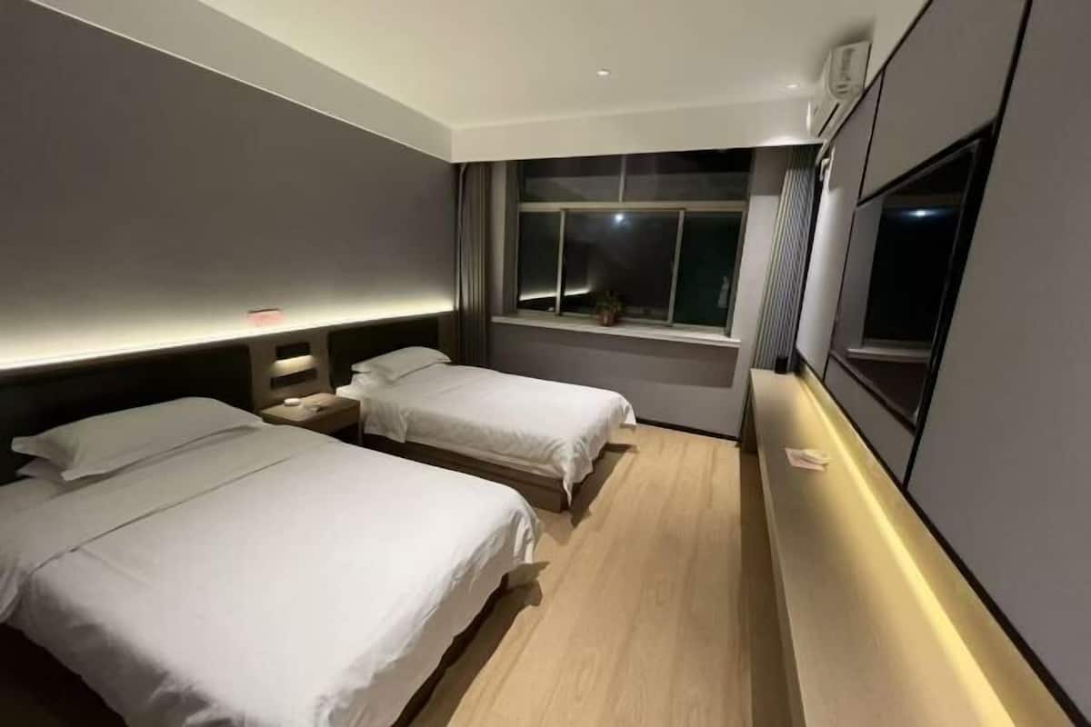 A hotel room with two beds, a window with curtains, a bedside table, and a wall-mounted TV.