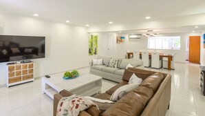 Ein modernes Wohnzimmer mit einem großen Flachbildfernseher, einem Couchtisch mit Dekoration, einem braunen Ledersofa und einem Essbereich mit weißem Tisch und Stühlen.