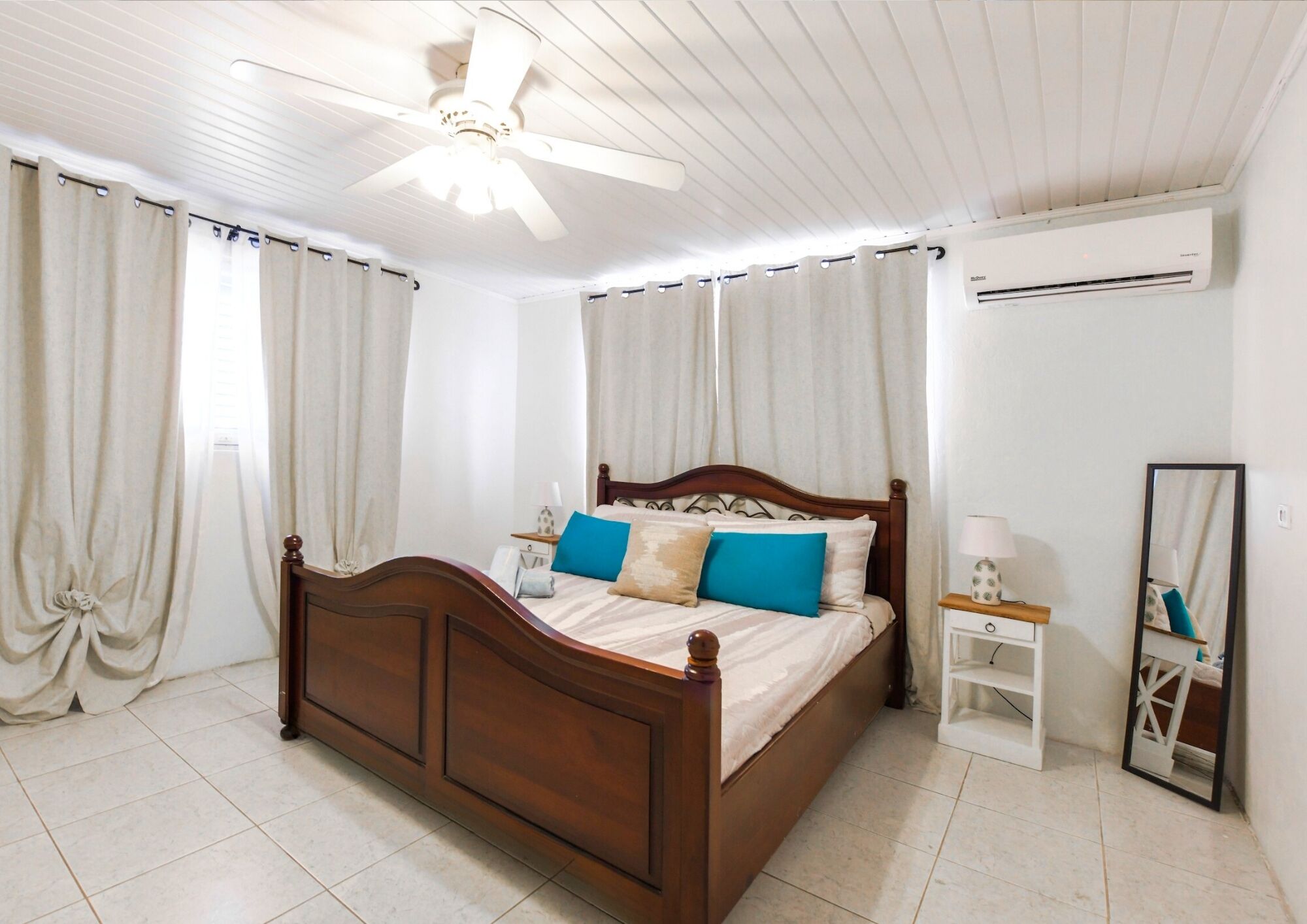 A bedroom with a wooden bed, white curtains, a ceiling fan, an air conditioning unit, a bedside table with a lamp, and a mirror.