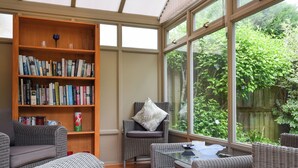 A cozy reading nook with wicker furniture, a large bookshelf, and a view of greenery through large windows. - 2 bedroom accommodation in Eynsham (Eynsham)