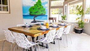 Un comedor con una mesa de madera, sillas blancas y un cuadro de un árbol junto al mar.