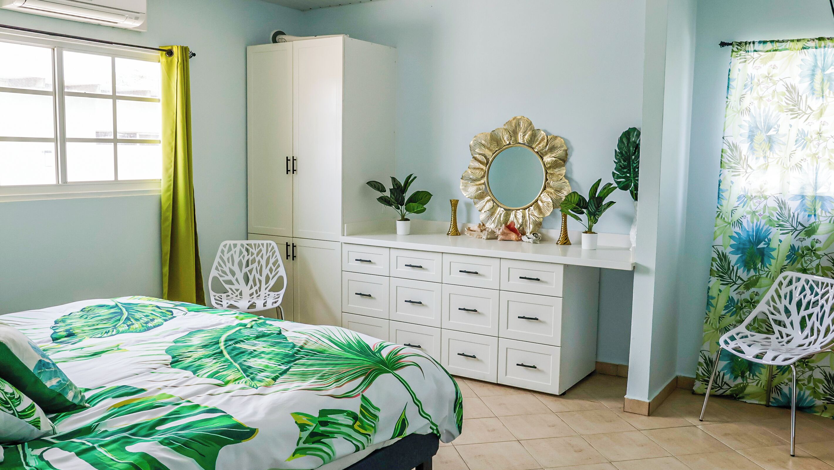 A bedroom with a bed, a white wardrobe, a dresser, two chairs, and a window with curtains.