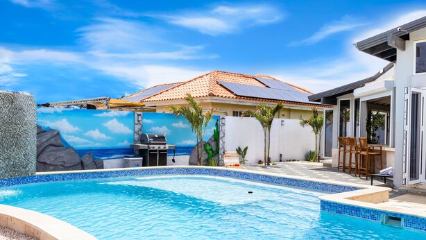 Piscina con un mural de una escena de playa, zona de barbacoa y patio con sillas.