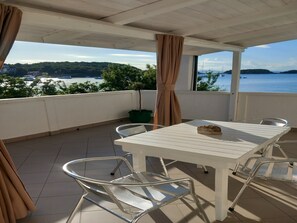 Ein Balkon mit weißem Tisch und Stühlen, einem Blumentopf und Blick auf das Meer und die Hügel.