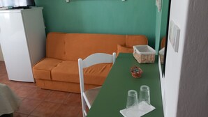A room with a green wall, a white sofa, a white table with a green tablecloth, a white chair, a small basket, and a television on a white cabinet.