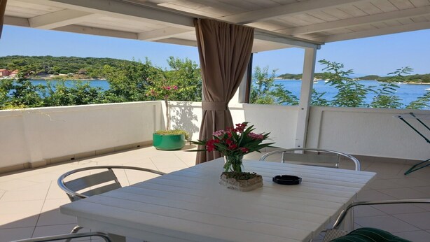 A covered outdoor seating area with a table and chairs, a potted plant, and a view of the sea and greenery.
