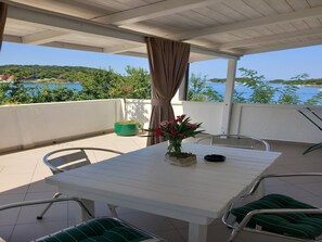 Ein überdachter Außenbereich mit Tisch, Stühlen, einer Topfpflanze und Blick auf Meer und Grün.