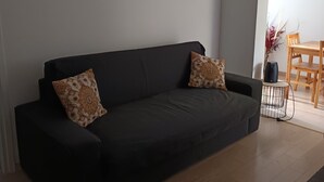 Un sofá de color oscuro con cojines estampados, una mesita auxiliar con un altavoz y una mesa de comedor de madera con sillas.