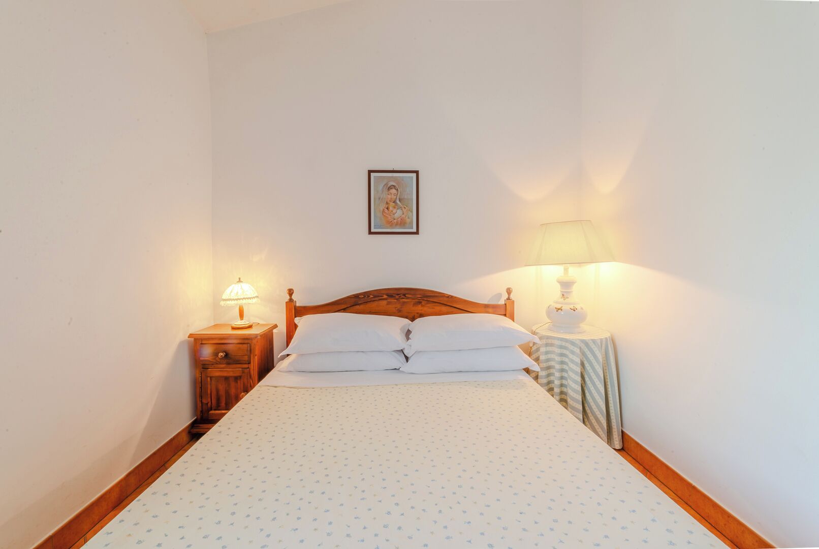 Un dormitorio con una cama de madera, dos mesitas de noche con lámparas, un cuadro en la pared y una colcha estampada.